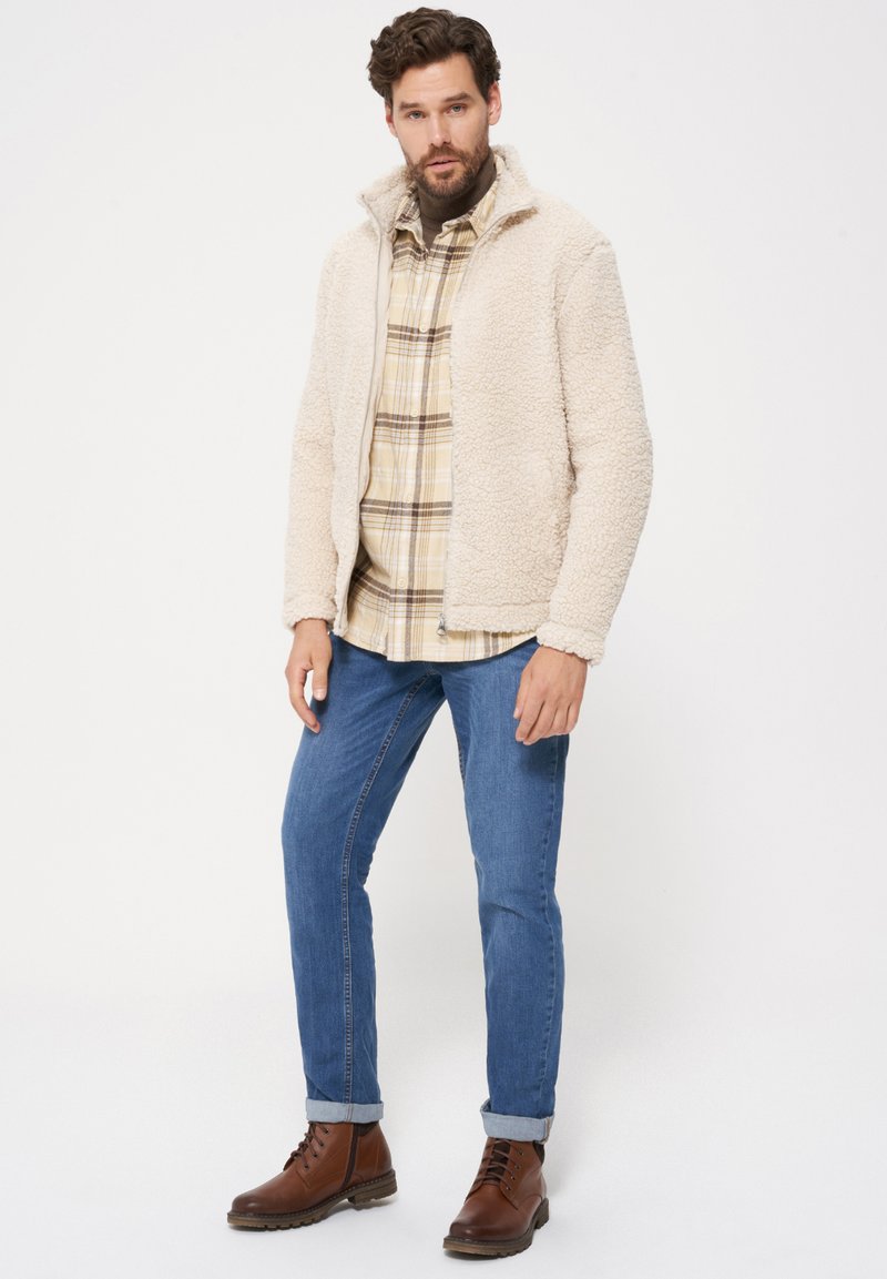 Beżowa polarowa kurtka z wysokim kołnierzem, noszona na koszuli w kratę. Model połączył ją z niebieskimi dżinsami i brązowymi, sznurowanymi botkami. Prosty, casualowy design.