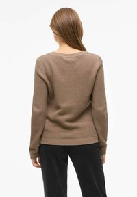 Maglione marrone con texture, collo rotondo e orlo a coste. Indossato con pantaloni neri, mostra una vestibilità rilassata e un motivo lavorato a maglia sul tessuto.