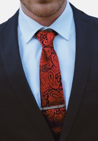 Corbata de paisley roja y negra con textura, que lleva un clip de corbata plateado y se usa sobre una camisa de vestir azul clara y una chaqueta de traje negra.