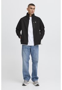 Zwarte zip-up jack met een staande kraag, een voorvak en zijzakken. Draag het over een wit T-shirt en combineer het met blauwe spijkerbroeken en sneakers.