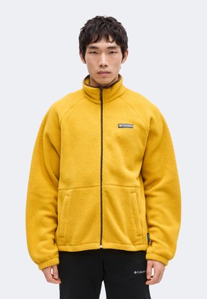 Jeune homme portant une veste polaire jaune moutarde avec fermeture éclair à l'avant et logo Columbia, debout devant un fond clair uni.