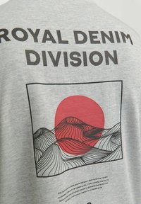 Γκρίζο ύφασμα με μαύρο κείμενο "ROYAL DENIM DIVISION" πάνω από ένα αφηρημένο κυματιστό ορεινό σχέδιο και έναν μεγάλο κόκκινο ήλιο σε τετράγωνο πλαίσιο.