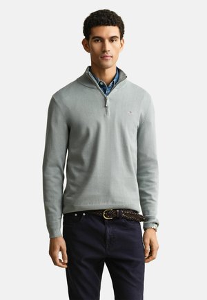 Mann trägt hellgrauen Pullover mit Viertelreißverschluss über blauem Hemd mit Kragen, dunkle Hose mit geflochtenem Gürtel, steht vor einfachem hellem Hintergrund.