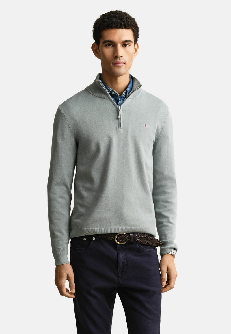 Homme portant un pull gris clair à quart de fermeture éclair sur une chemise bleue à col, pantalon foncé avec ceinture tressée, debout sur un fond clair uni.