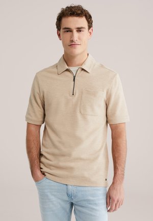 Beige poloshirt met korte mouwen, ritssluiting bij de hals en een borstzak. Getextureerde stof, losse pasvorm, gecombineerd met lichtblauwe jeans.