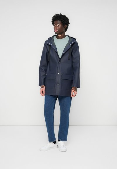 Marineblauer wasserdichter Mantel mit Kapuze, ausgestattet mit zwei vorderen Taschen, Druckknopfverschlüssen und einem kontrastierenden grünen Innenfutter. Kombiniert mit blauen Jeans und weißen Sneakern.