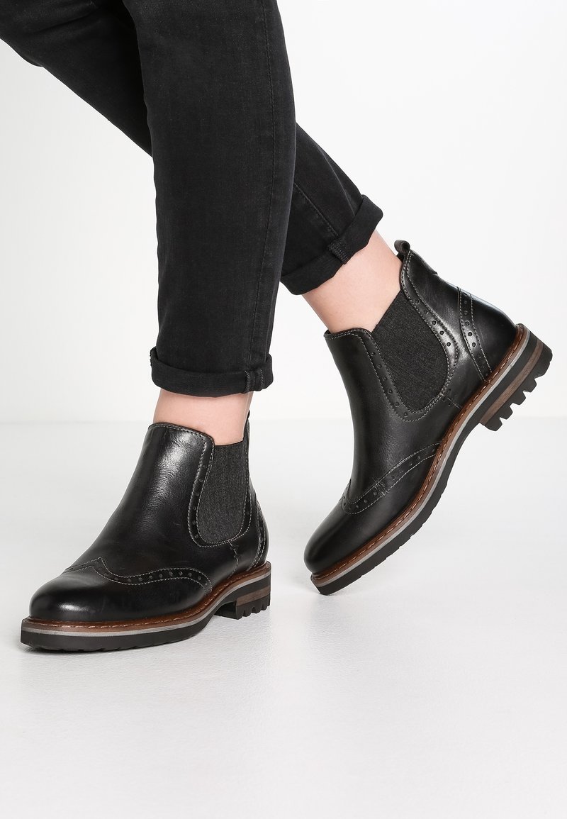 Bottes Chelsea en cuir noir avec panneaux élastiques sur les côtés, détails brogue et semelle texturée. Le talon est légèrement empilé pour un peu plus de hauteur.