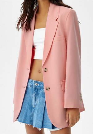Femme portant un blazer rose par-dessus un crop top blanc et une mini-jupe plissée en jean bleu avec un ourlet effiloché.