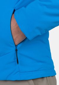 Sininen takki, jossa on sivussa vetoketjullinen tasku, valmistettu sileästä kankaasta, muotoon ommeltu ja jossa on siisti ompelu.