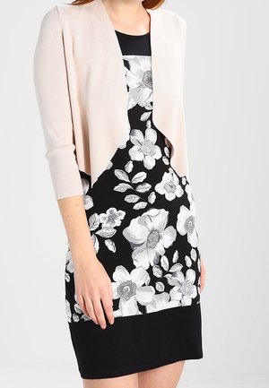 Femme portant une robe noire à grand imprimé floral blanc et un cardigan court rose clair ouvert devant avec des manches trois-quarts.