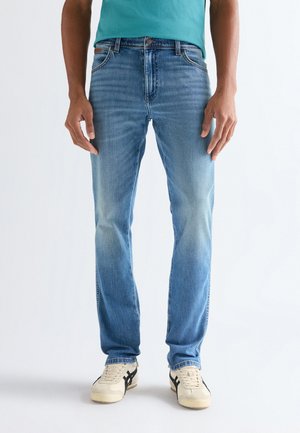 Férfi világoskék egyenes szárú farmerben, türkiz ingben és fekete csíkkal díszített fehér sportcipőben, fehér háttér előtt állva.