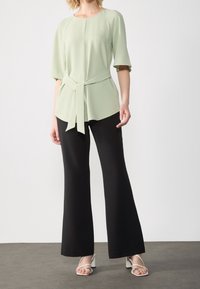 Haut vert clair à manches courtes avec un lien à la taille, associé à un pantalon noir à jambes larges. Le mannequin porte des sandales blanches à talons.