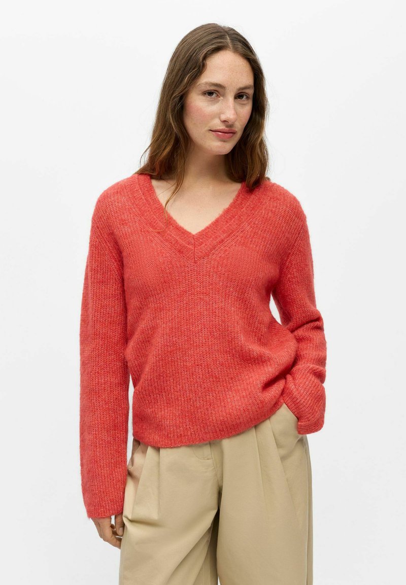 Femme portant un pull corail rouge à col en V et un pantalon beige taille haute à jambes larges sur un fond blanc uni.