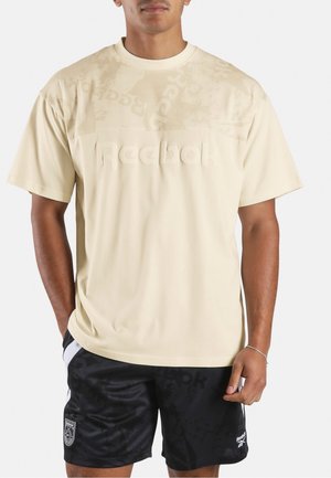 Mann trägt beige Reebok T-Shirt und schwarze Reebok Shorts mit Logodetails, steht mit entspannten Händen vor einem schlichten Hintergrund.