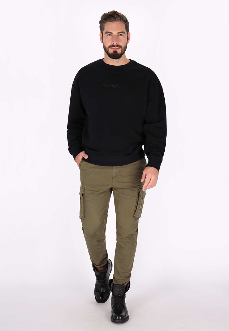 Ein bärtiger Mann geht vorwärts und trägt einen schwarzen Sweatshirt, olivfarbene Cargohosen und schwarze Stiefel vor einem schlichten hellen Hintergrund.