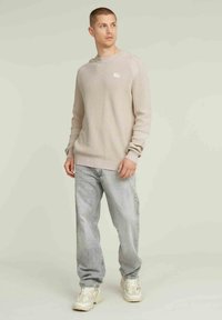 Maglione a coste beige chiaro con scollo rotondo e un piccolo logo. Abbinato a jeans dritti grigio chiaro e sneakers beige.