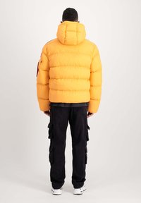 Chaqueta de plumas amarilla con capucha, textura acolchada y bolsillos con cremallera. Combinada con pantalones cargo negros que tienen múltiples bolsillos laterales.