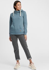 Sudadera azul con capucha, cordones crema, bolso frontal y un pequeño parche de logo marrón. Combinada con jeans grises remangados y zapatillas crema.