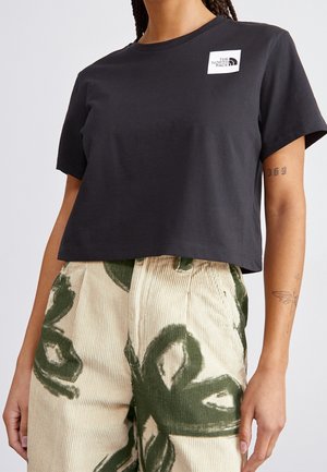 Kobieta ubrana w czarny krótki T-shirt z logo The North Face i kremowe spodnie ze sztruksu z zielonymi abstrakcyjnymi wzorami.