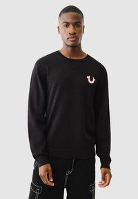 Schwarzer gestrickter Pullover mit Rundhalsausschnitt, langen Ärmeln und einem kleinen roten Logo auf der linken Brust. Das Material wirkt weich mit einer glatten Textur.