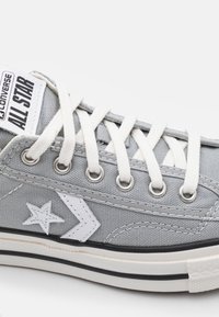 Sneaker Converse All Star gris avec lacets blancs, logo étoile et chevron blancs, et semelle en caoutchouc blanche à rayures noires.