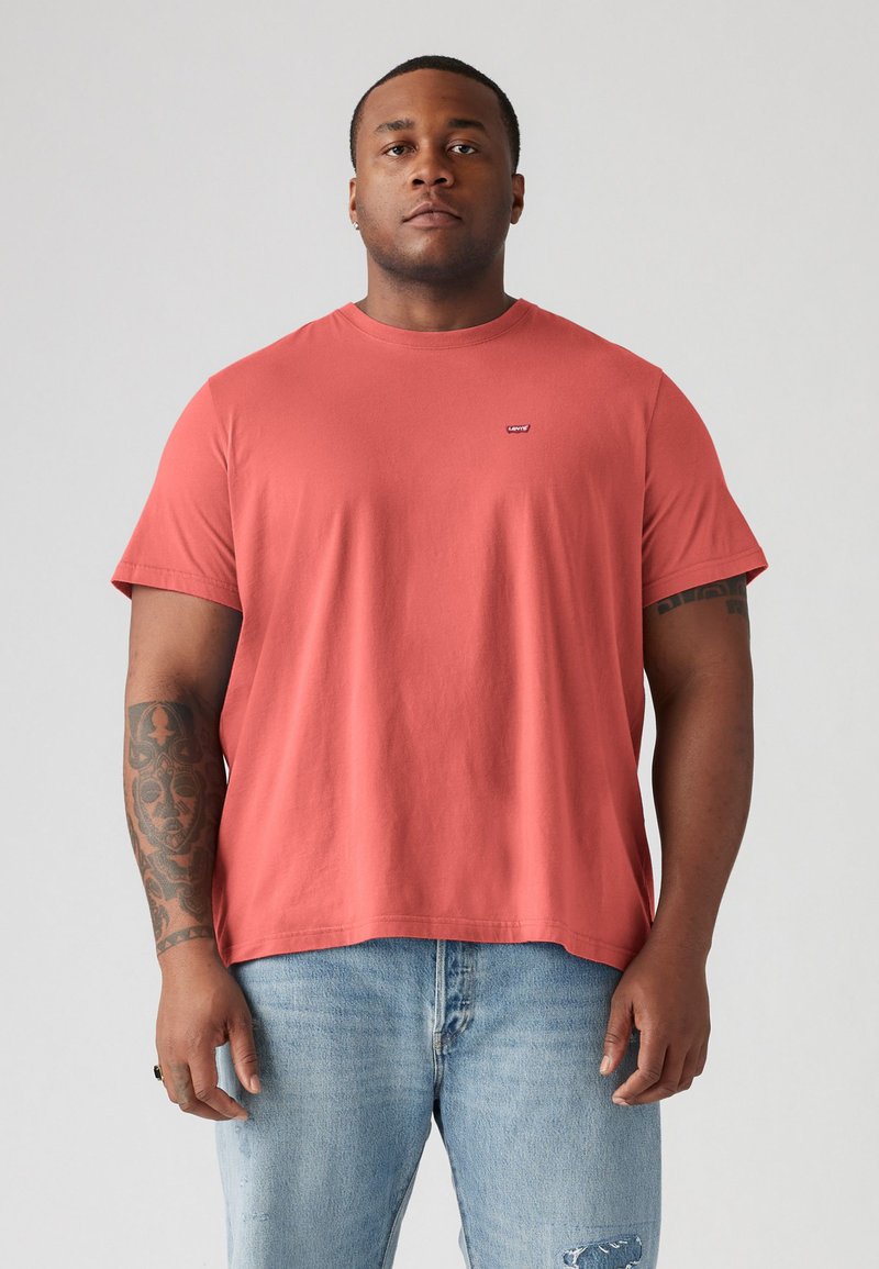 Koralfarvet bomulds t-shirt med rund hals og korte ærmer, der har et lille logo på brystet. Båret sammen med lyseblå slidte jeans.