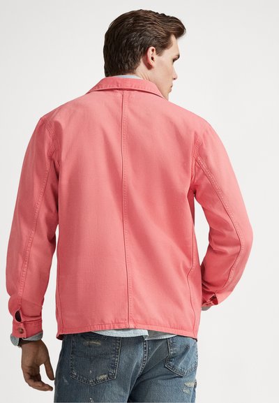 Vīrietis, kurš nēsā izbalējušu rozā jaku pār gaiši zilu kreklu un nodilušas zilās džinsas, stāv ar muguru pret vienmērīgi baltu fonu.