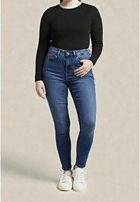 Kobieta ubrana w niebieskie, dopasowane dżinsy z wysokim stanem, czarną bluzkę z długim rękawem i białe trampki, stojąca na tle jednolitego beżowego tła.