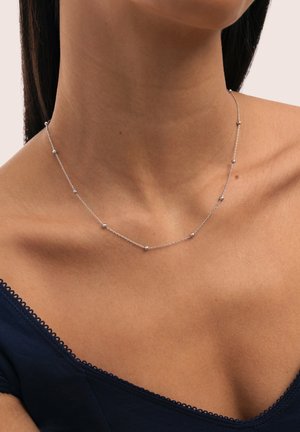Collar delicado de cadena de plata con pequeñas cuentas redondas repartidas de manera uniforme, llevado en el cuello y la clavícula de una mujer, con un top azul marino de hombros descubiertos visible.