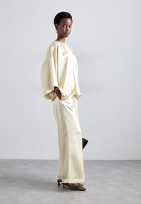 Haut en satin crème à manches larges, associé à un pantalon large assorti. Des chaussures à talons sombres et un sac noir complètent la tenue.