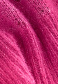 Rosa stickat tyg med mjuk, luddig textur. Har vertikal ribbning och lösa trådar, vilket visar en rik, livfull färg.