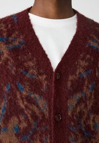 Cardigan bordeaux avec une texture duveteuse, col en V et un motif abstrait marron-bleu. Fermé par deux boutons foncés à l'avant.