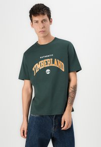 Camiseta de algodón verde con "TIMBERLAND" en texto naranja en negrita, con cuello redondo y mangas cortas. Llevada con pantalones vaqueros de denim azul.