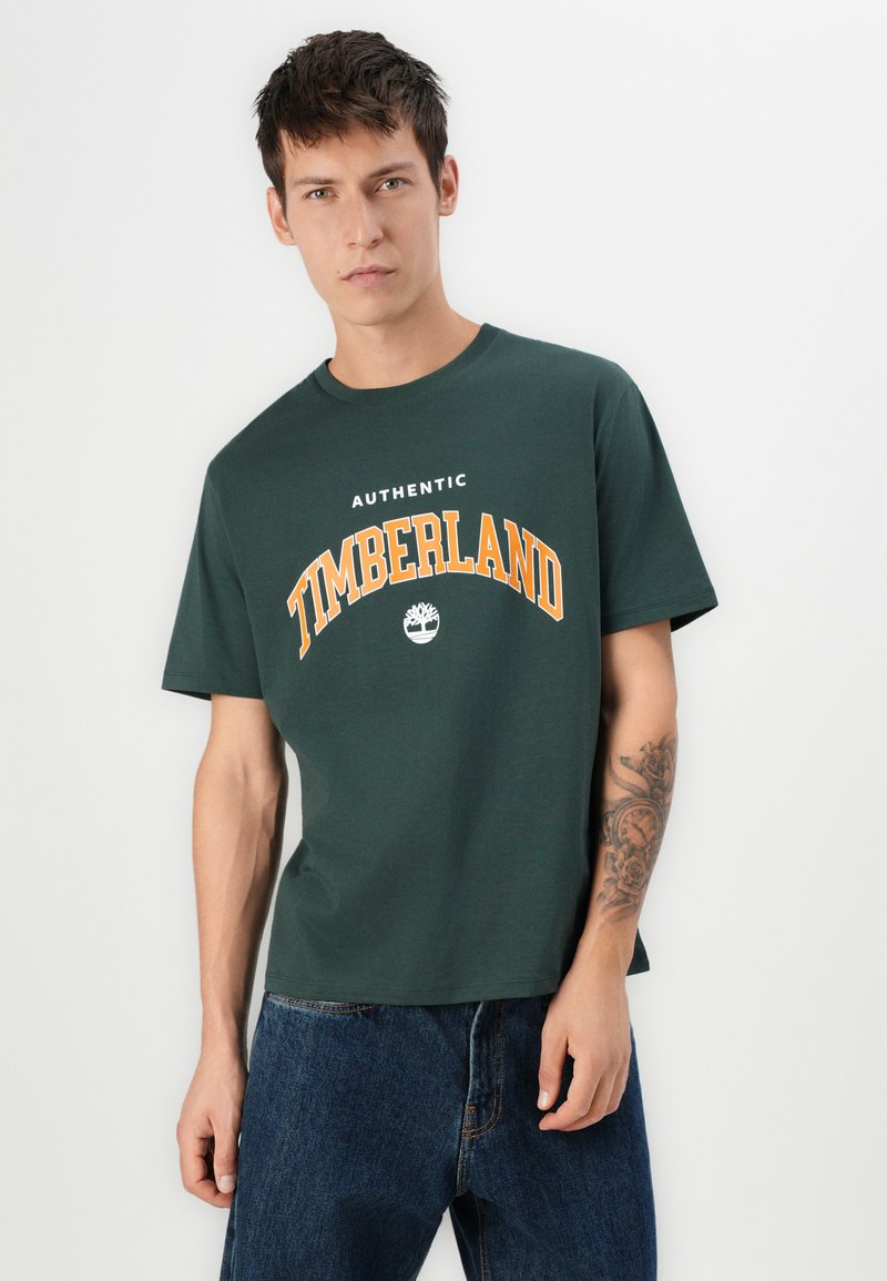 T-shirt verde in cotone con la scritta "TIMBERLAND" in grassetto arancione, scollatura tonda e maniche corte. Indossato con jeans di denim blu.