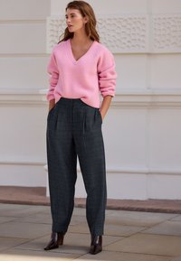 Skarp pink ribstrikket sweater med V-udskæring, kombineret med skræddersyede mørkegrå bukser med et diskret ternet mønster. Brune sko med hæl.