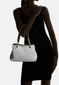 Kobieta w czarnej sukience bez rękawów trzyma jasnoszary torebkę z podwójnymi czarnymi uchwytami na lewym ramieniu.