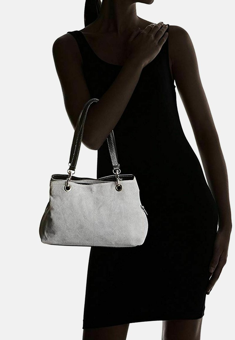 Kobieta w czarnej sukience bez rękawów trzyma jasnoszary torebkę z podwójnymi czarnymi uchwytami na lewym ramieniu.
