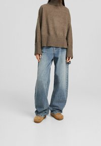 Brązowy sweter z golfem z dzianiny z prążkowanym dołem, w połączeniu z jasnoniebieskimi, luźnymi dżinsami i brązowymi butami wsuwanymi na jednolitym tle.