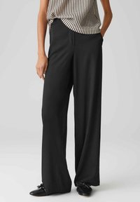 Schwarze, weit geschnittene Hose aus glattem Stoff, mit gefaltetem Vorderteil und Eingrifftaschen. Getragen mit einem leichten, strukturierten Oberteil und schwarzen Schuhen.