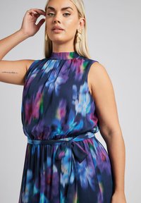 Abito senza maniche in tessuto fluido con un motivo floreale multicolore su uno sfondo scuro, dotato di un collo alto e vita legata.