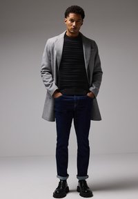 Grauener Wollmantel über einem schwarzen strukturierten Pullover, kombiniert mit dunkelblauen Jeans und schwarzen klobigen Stiefeln. Einfaches Design und neutrale Farben.
