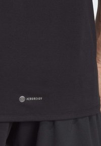 Tissu noir avec logo blanc "AEROREADY" et icône de globe près de l'ourlet, porté par-dessus un short ou une jupe texturée foncée sur le côté d'une personne.