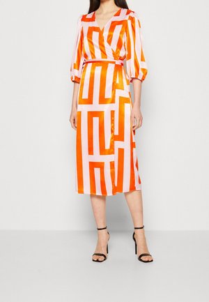 Frau trägt ein Midi-Wickelkleid mit orangefarbenen geometrischen Streifen und voluminösen Dreiviertelärmeln, kombiniert mit schwarzen Pumps mit Knöchelriemen.