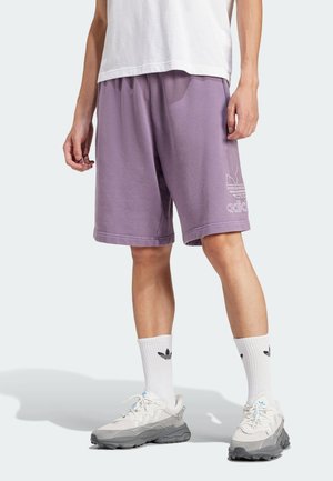 Osoba nosząca fioletowe spodenki Adidas, białe skarpetki Adidas, białe i szare sneakersy oraz białą koszulkę, stojąca na jasnym tle.