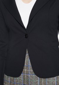 Blazer noir en tissu texturé, avec un col cranté, fermeture à un bouton, et deux poches avant, porté sur une jupe à motifs.