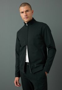 Schwarze Zip-Jacke mit hohem Kragen, mit glatter Textur, Seitentaschen und eng anliegenden Ärmeln, über einem weißen Hemd getragen.