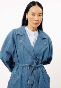 Jeans trenchcoat met grote revers, zijzakken en een ceintuur in de taille. Heeft een relaxte pasvorm en contrasterende stikdetails.