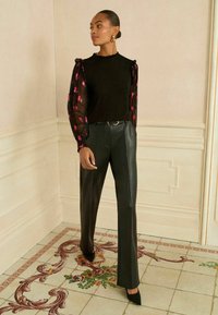 Fekete felső áttetsző, rózsaszín mintás ujjal, magas derekú fekete bőrnadrággal és hegyes orrú fekete sarkú cipővel párosítva. Egyszínű háttér.