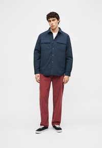 Νέος άνδρας που φοράει μπλε σακάκι-πουκάμισο με κουμπιά, λευκό εσώρουχο, κόκκινο παντελόνι και μαύρα αθλητικά παπούτσια, στέκεται μπροστά σε ένα απλό ανοιχτό φόντο.