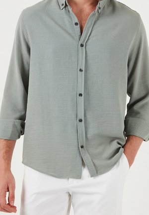 Mies pukeutuneena vaaleanvihreään pitkähihainen nappipaita, hihat käärittyinä, ja valkoiset housut, toinen käsi taskussa, yksiväristä taustaa vasten.